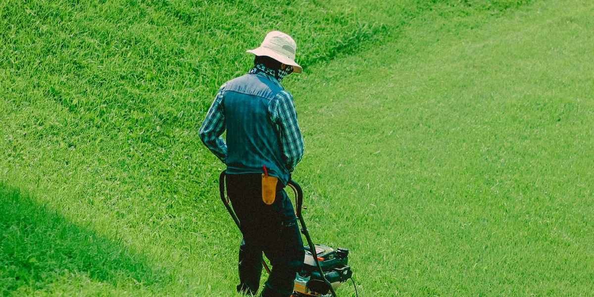 A person in a wide-brimmed sun hat pushing a mower across a lush green lawn, a freshly cut stripe trailing behind them